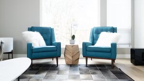 A modern living room with two blue armchairs, a white fluffy pillow, a wooden side table, and a view of trees through large windows. - Capitol Hill Townhouse with Private Rooftop Deck — Space Needle & Skyline Views (Seattle)