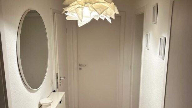 Un corridoio con pavimento in legno, pareti bianche, uno specchio rotondo, un mobile da trucco bianco con una ciotola e un particolare lampadario.