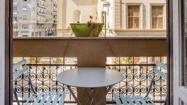 Ein Balkon mit einem runden Tisch und zwei blauen Stühlen, einem grünen Pflanzgefäß mit Pflanzen und Blick auf ein Gebäude mit vielen Fenstern.