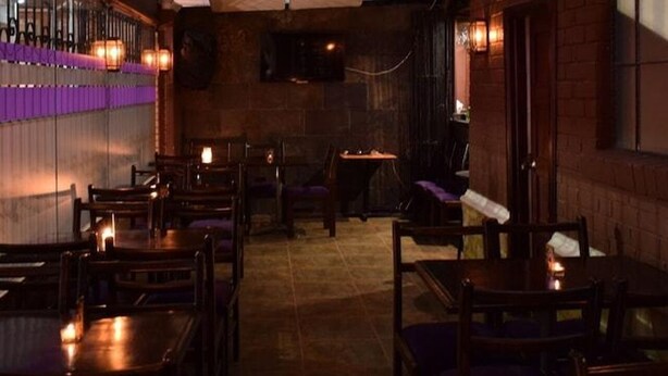 A dimly lit bar area with wooden tables and chairs, a brick wall, and a television mounted on the wall. - Hotel Casa Morada (Bogotá)