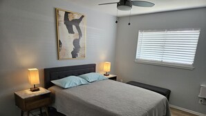 Une chambre à coucher comprenant un lit, deux lampes de chevet, un ventilateur de plafond, une fenêtre avec des stores et une œuvre d’art accrochée au mur.