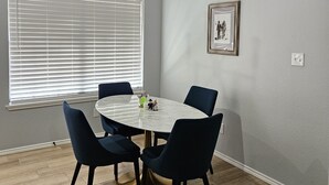 Un coin repas avec une table ronde, des chaises foncées, un lustre, une fenêtre avec des stores et un tableau encadré au mur.