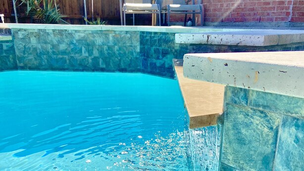 A swimming pool with clear blue water, a stone wall, and a wooden fence in the background. - Spa & Pool + Stay in Harry Potter Theme Home (Frisco)