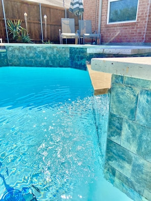 A swimming pool with clear blue water, a stone wall, and a wooden fence in the background.