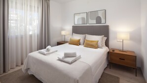 Una cama bien hecha con sábanas blancas y dos almohadas, una mesita de noche con lámpara y una ventana con cortinas traslúcidas.