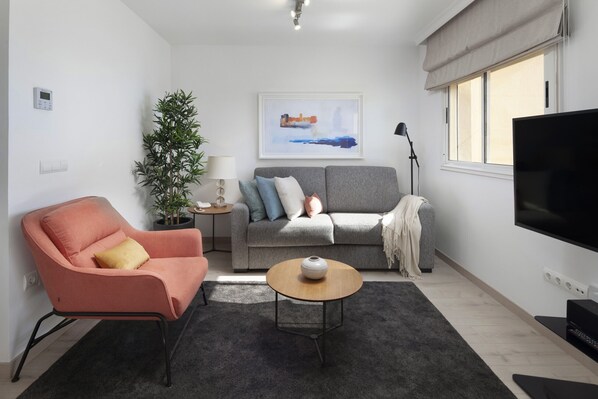 Una sala de estar moderna con un sofá gris, un sillón rosa, una mesa de centro, un televisor y un amplio ventanal con cortinas.