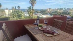 A wooden table with a bottle of wine, a glass, and a plate of food on a balcony overlooking a residential area with palm trees and buildings.