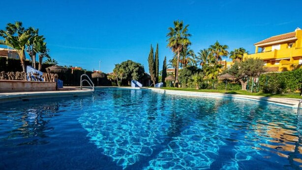 A clear blue pool surrounded by palm trees and a yellow building.