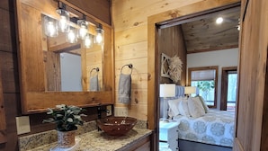 A wooden cabin interior with a kitchen area featuring a granite countertop, a large mirror, and a bowl sink. A bedroom with a bed and a window is visible through an open door. - Incredible firepit, luxurious cabin, indoor&outdoor fireplaces. Minutes to town. (Morganton)
