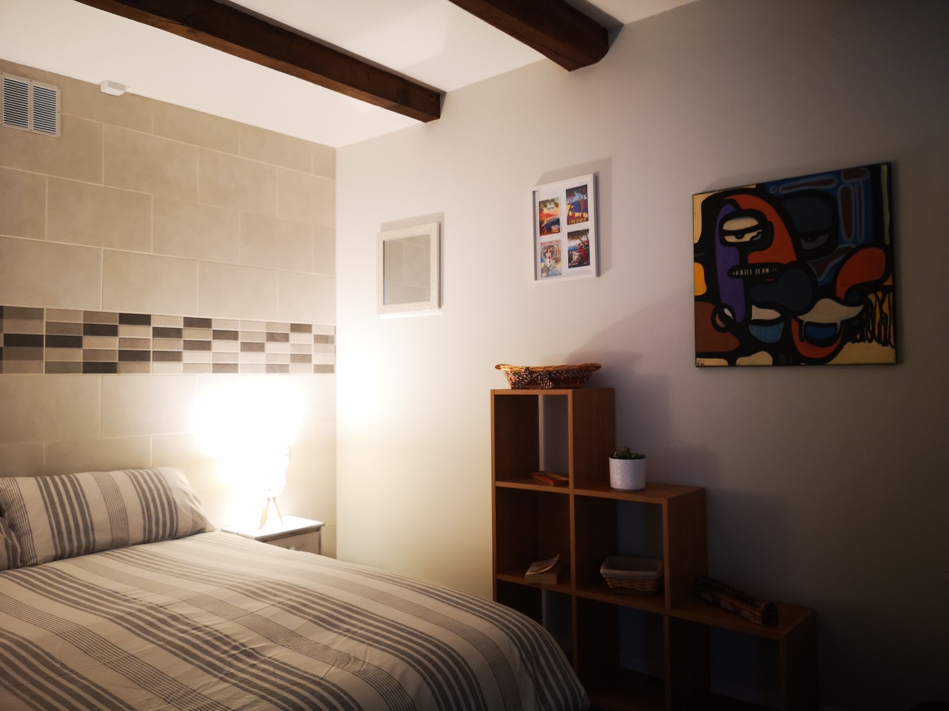 Ein Schlafzimmer mit einem Bett, einem Nachttisch, einem Regal mit Büchern und einem Korb, einer Wand mit Fliesen und einem gerahmten Bild.