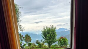 A room with a large window offering a view of mountains and greenery. There is a small round table and two green chairs. - Haut Monde King's Paradise Resort  (Tehri)