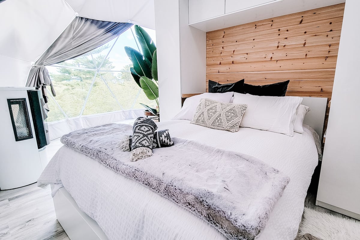 Ein modernes Schlafzimmer mit einem Holzkopfende, einem großen Fenster mit Vorhängen, einem Bett mit weißer Bettwäsche und einer kleinen Klimaanlage.