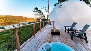 Una terraza de madera con un jacuzzi, sillones de descanso y una estructura en forma de domo geodésico, con vistas a un lago y un bosque.