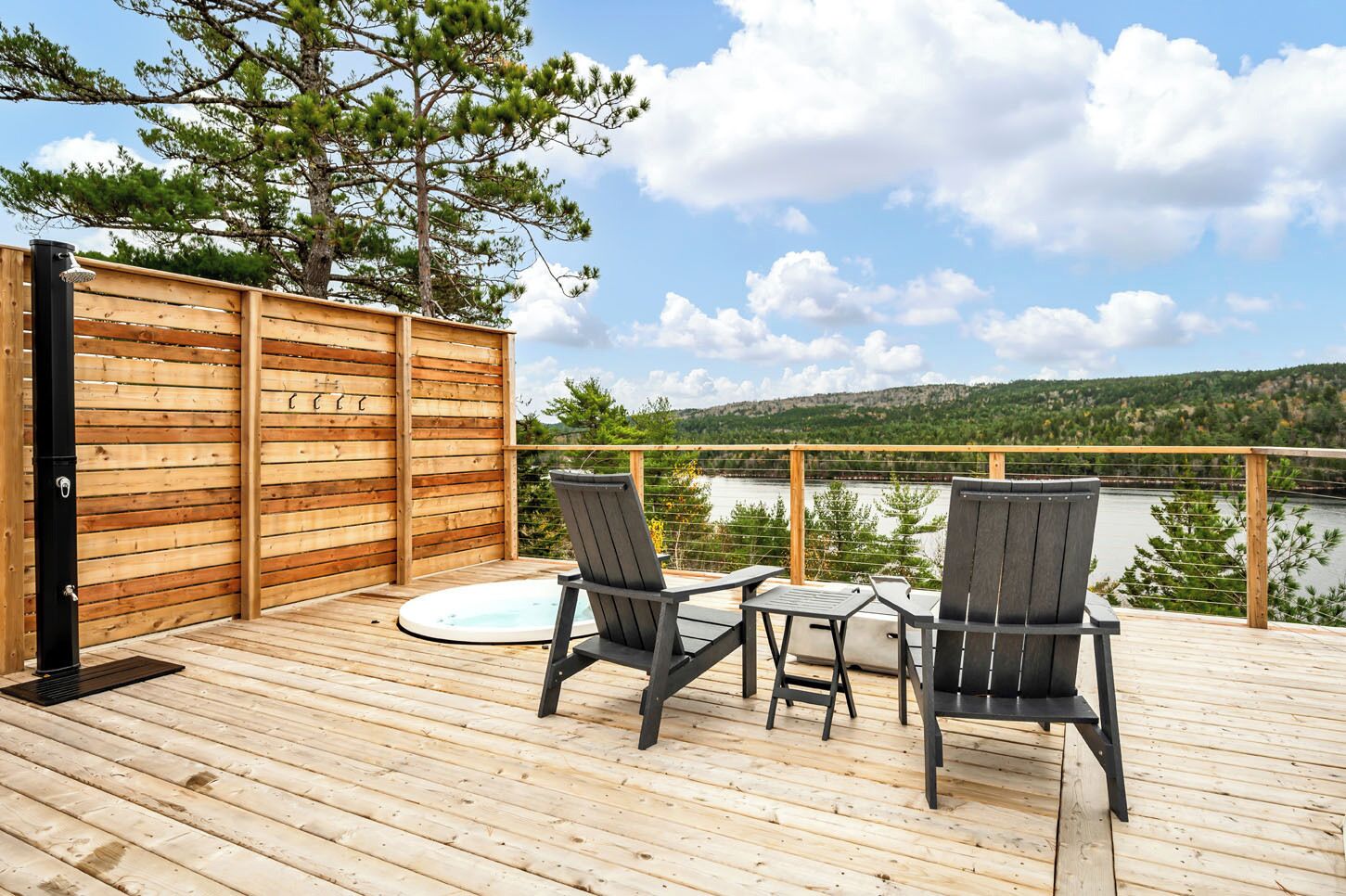 Una terraza de madera con dos sillas y un jacuzzi, con vistas a un lago y colinas boscosas.