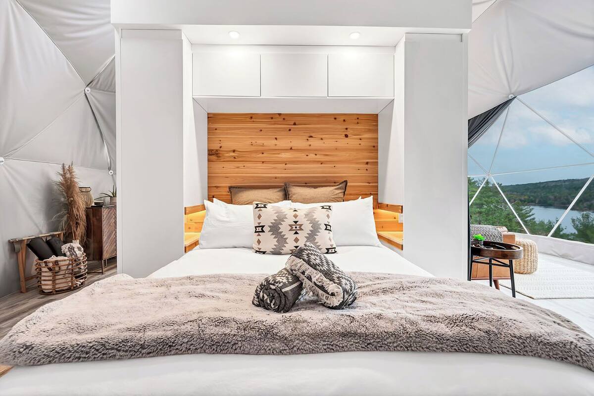 Ein modernes Schlafzimmer mit einem Holz-Kopfteil, einem großen Bett mit gemusterten Kissen, einem flauschigen Teppich und Blick ins Freie durch ein großes Fenster.