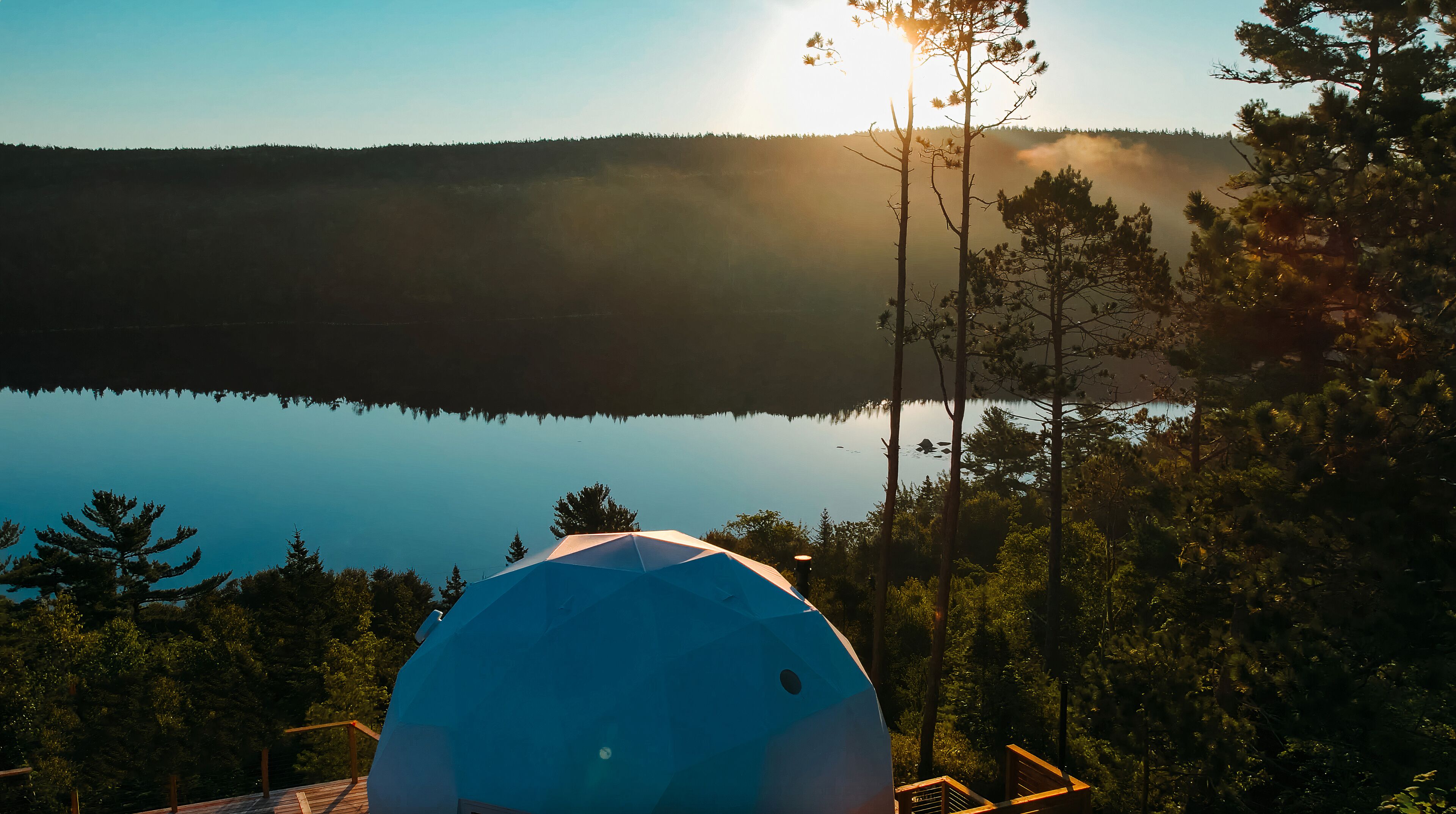 A dome-shaped tent with a clear view of a calm lake and forested hills in the background.