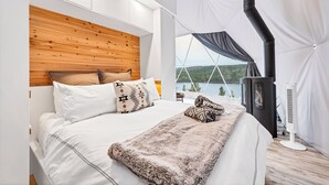 Une chambre moderne avec une tête de lit en bois, un grand lit avec une literie blanche, un plaid moelleux et une vue sur un lac, visible à travers une structure de fenêtre aux motifs géométriques.