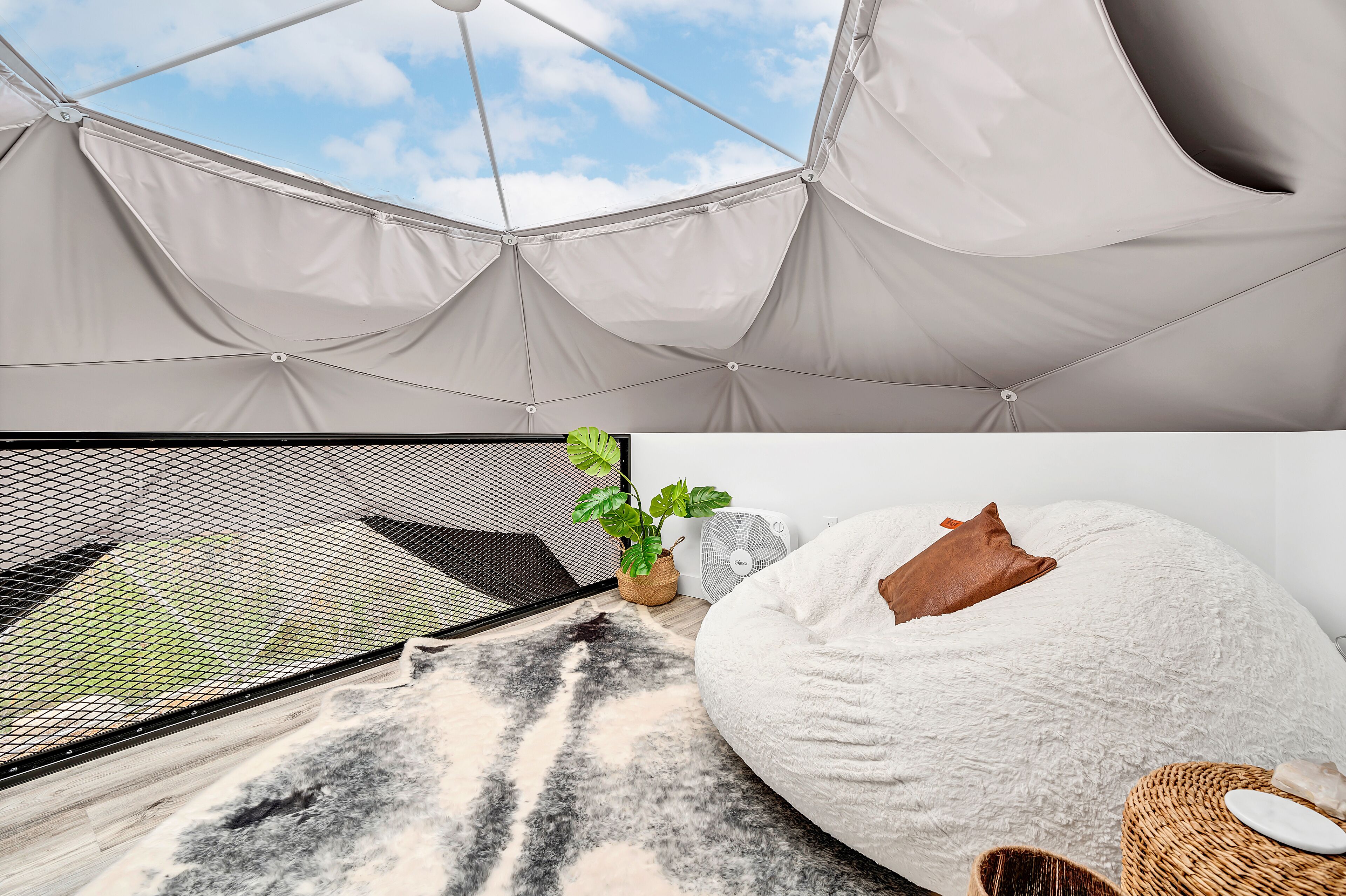 A tented room with a view of the sky, a bed with a pillow, a fan, a plant, and a woven basket.