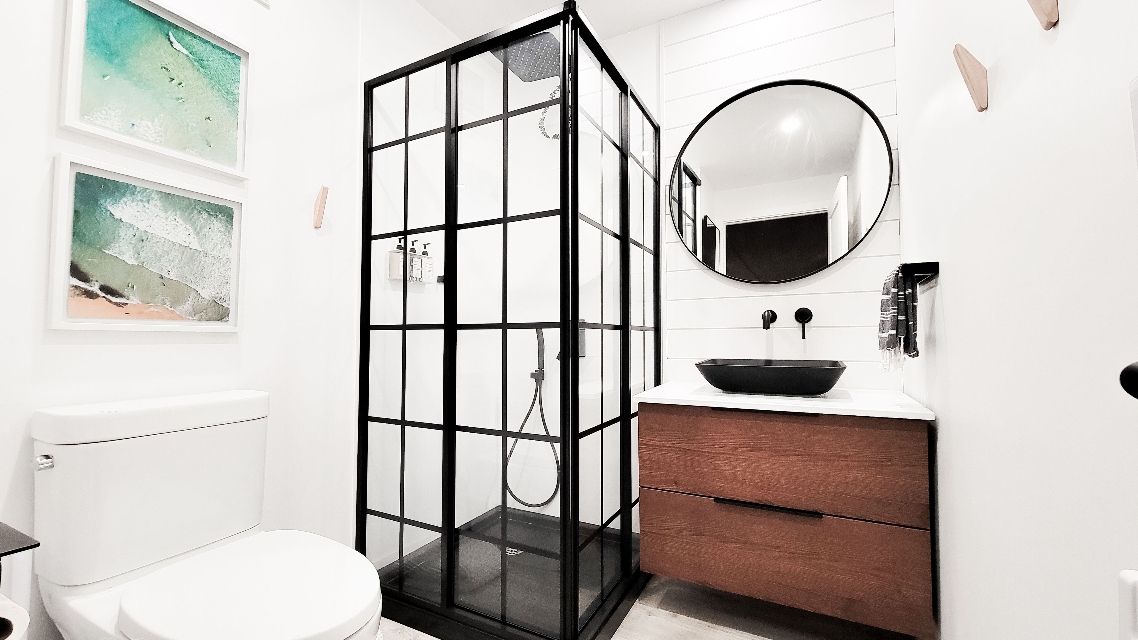 A modern bathroom with a glass-enclosed shower, a white toilet, a wooden vanity with a black sink, and a round mirror.