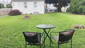 Un espacio al aire libre con césped, una mesa redonda y dos sillas, un edificio blanco al fondo y un árbol.