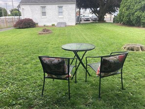 Un espace extérieur verdoyant avec une table ronde et deux chaises, un bâtiment blanc en arrière-plan et un arbre.