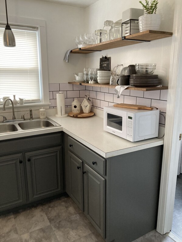 Une cuisine moderne avec des meubles gris, un plan de travail blanc, un four à micro-ondes encastré et une fenêtre avec des stores.