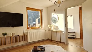 Een moderne woonkamer met een grote televisie, een houten radiator, een ronde tafel en uitzicht op een besneeerd landschap door het raam.