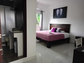 Ein Schlafzimmer mit Bett, Kopfstütze, Nachttisch, Fenster und einem Badezimmer mit Waschbecken und Toilette.