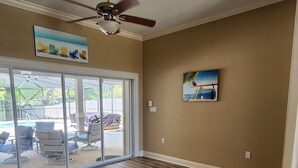 Una stanza spaziosa con ventilatore a soffitto, grandi porte scorrevoli in vetro che danno su un’area esterna con mobili da patio e un quadro appeso al muro.