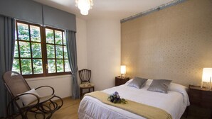 Een slaapkamer met een groot bed, twee nachtkastjes met lampen, een schommelstoel, een fauteuil en een raam met gordijnen.