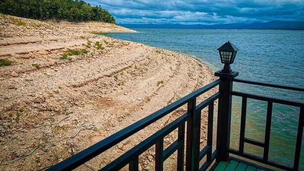 A view from a covered deck overlooking a rocky shoreline with a body of water and a forested area in the background. - Cozy, friendly, surroundings with nature ,Thai culture and  life style. (Nasuan Srisawat)