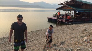 A man and a child near a dock with a boat, rocky shore, and mountains in the background. - Cozy, friendly, surroundings with nature ,Thai culture and  life style. (Nasuan Srisawat)