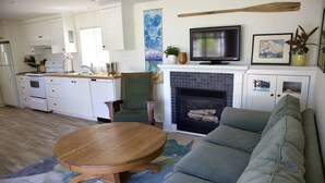 Une cuisine moderne avec des meubles blancs, une table à manger en bois et un canapé gris dans un salon doté d’une cheminée.