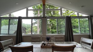 Un comedor con una mesa grande y sillas, ventanas amplias con cortinas y vistas a árboles y edificios.