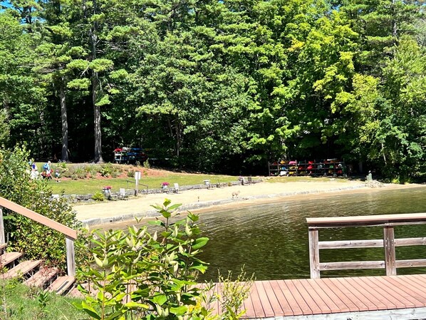 A wooden deck overlooking a calm lake, surrounded by dense trees and a grassy area with a few people and parked vehicles in the background.
