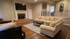 A spacious living room with a fireplace, a large sofa, a coffee table, and a TV mounted on the wall.