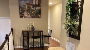 A hallway with a wooden floor, a bench, a dining table with chairs, a potted plant, and a painting on the wall.