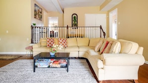 A living room with a beige sectional sofa, a coffee table with magazines, and a staircase with a railing.