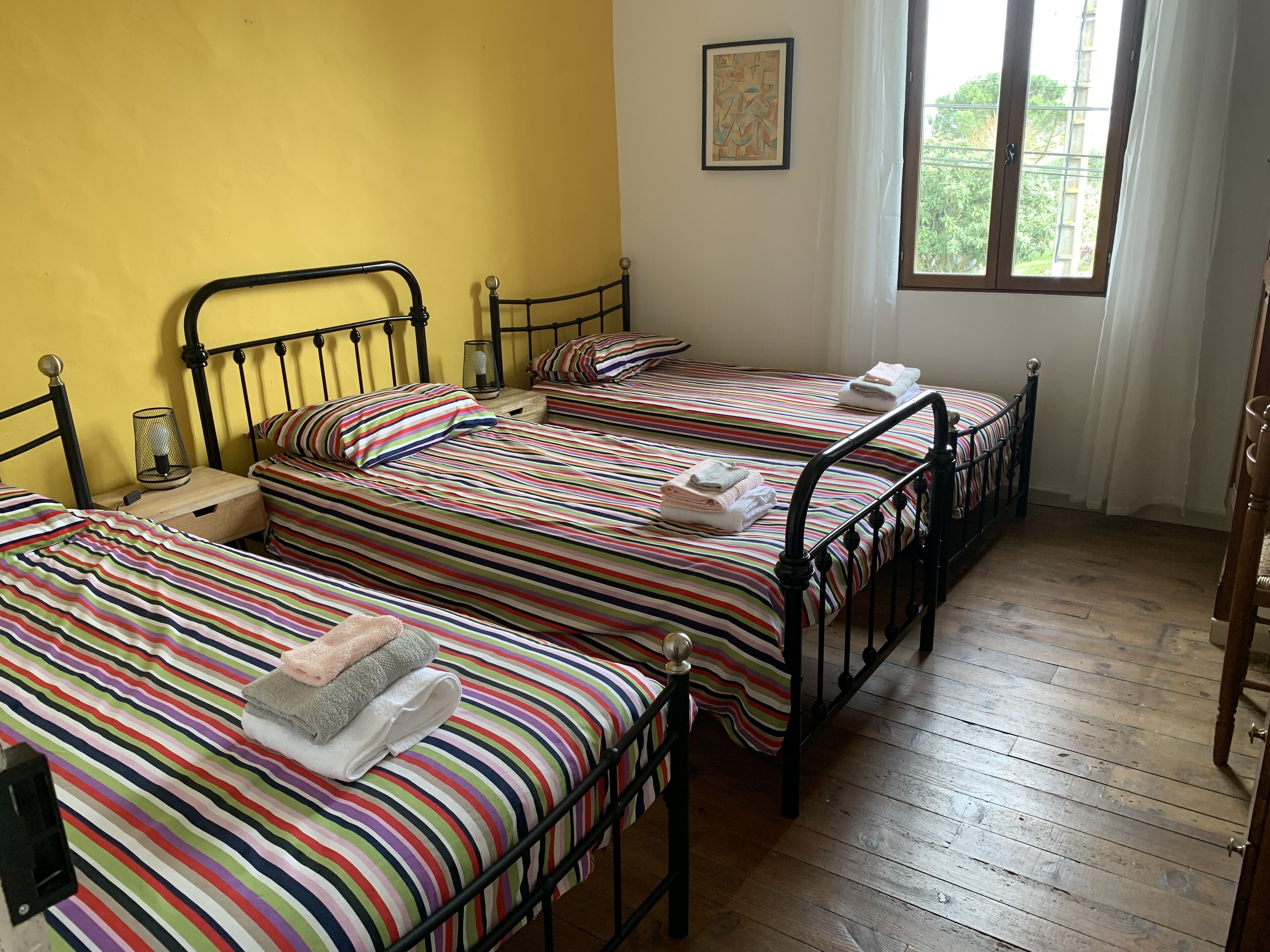A room with three beds, each with striped bedding and towels, a small table with a lamp, and a window with curtains.