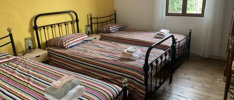 A room with three beds, each with striped bedding and towels, a small table with a lamp, and a window with curtains.
