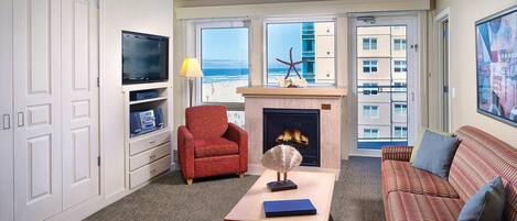 A living room with a fireplace, a television, a red armchair, a sofa, and a view of the ocean through large windows.