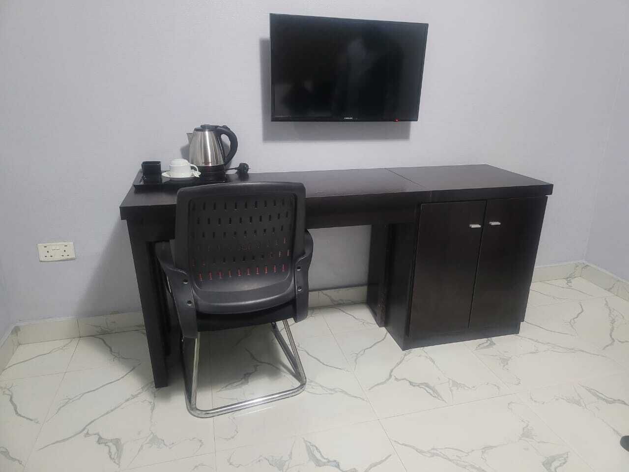 Een donker houten bureau met daarboven een televisie, een zwarte bureaustoel en een koffiezetapparaat op het bureau.