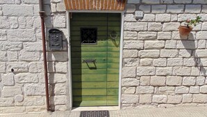 Un bâtiment en briques blanches avec une porte verte, une petite fenêtre ornée d’une grille décorative et un panneau indiquant le numéro « 64 » sur le mur.