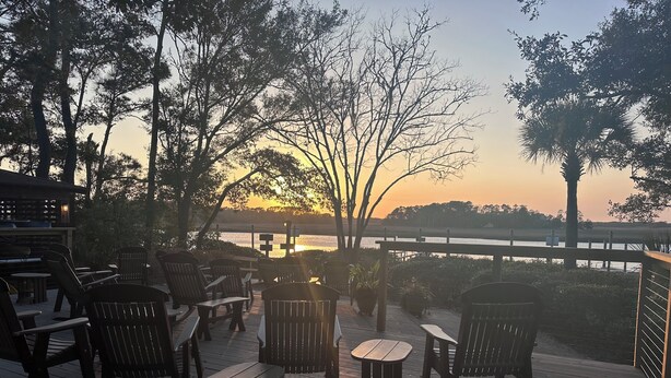 Une terrasse en bois avec des sièges extérieurs, offrant une vue sur un plan d’eau et sur le coucher du soleil.