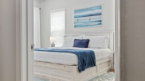 Un dormitorio con estructura de cama de madera, ropa de cama blanca, un plaid azul y un cuadro de una playa.