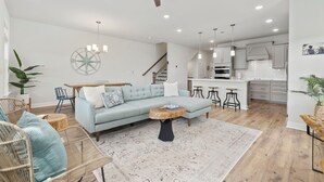 Un salón amplio con un sofá azul claro, una mesa de centro de madera y sillas de mimbre. Se ve en el fondo un comedor con mesa y sillas de madera. La cocina es moderna, con armarios blancos y una isla grande.