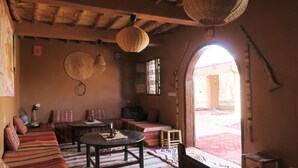 Une pièce chaleureuse avec un plafond bas, des paniers tressés suspendus aux poutres, une petite table avec une bougie, et une vue sur un espace extérieur, accessible par un passage en arc de cercle.