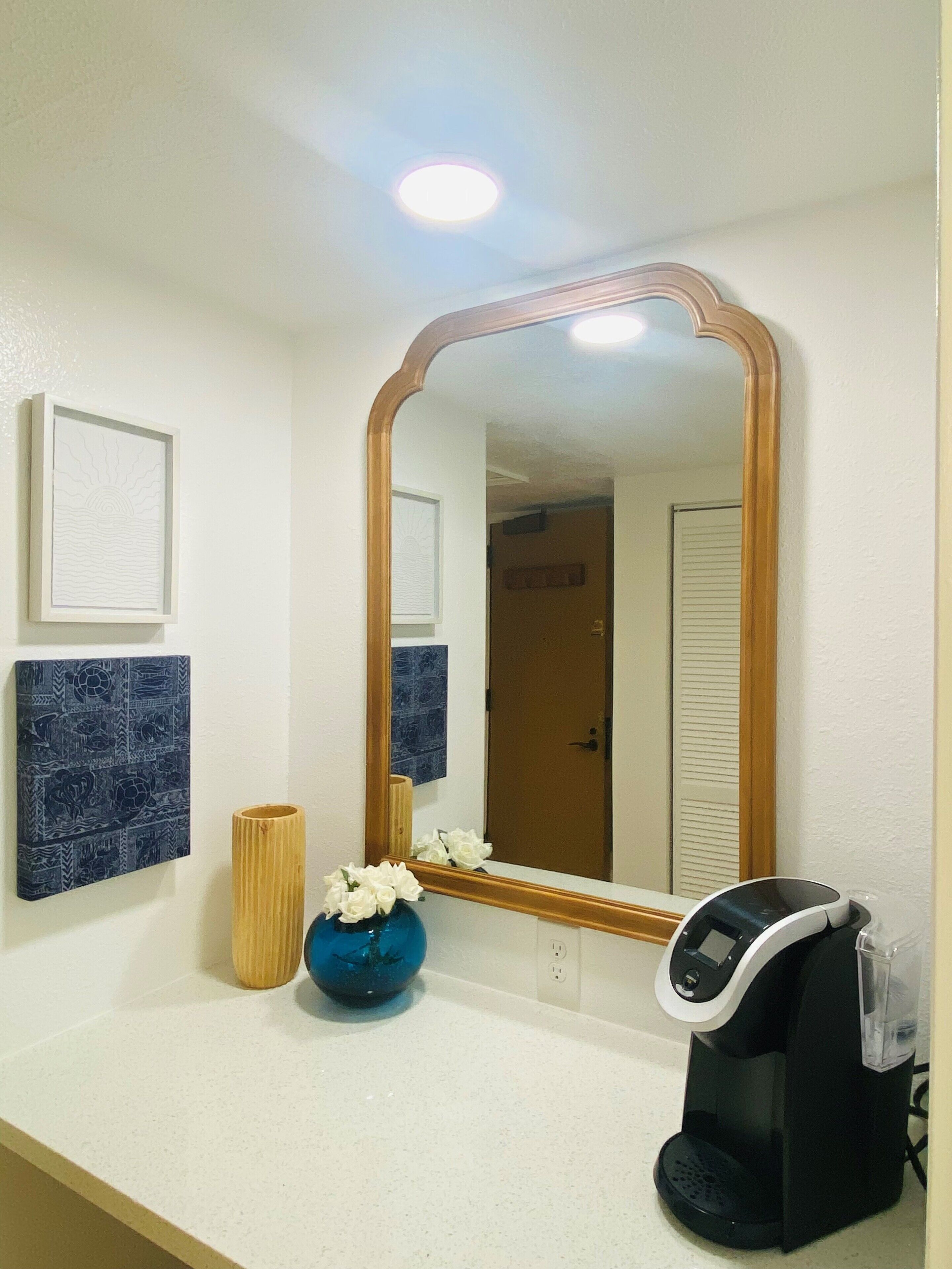 A bathroom with a white countertop, a large mirror, a coffee maker, a vase with white flowers, and two framed pictures on the wall.