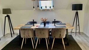 A modern dining area with a wooden table, three cushioned chairs, a vase of dried flowers, and pendant lighting.