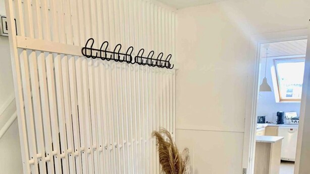 A room with a white partition, a bench, a vase with pampas grass, and a kitchen visible through an open door.